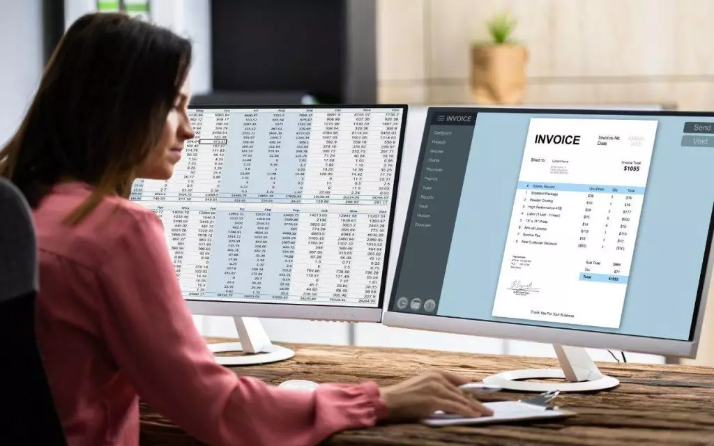 A businesswoman is sitting at a desk with two monitors in front of her, diligently tracking expenses.