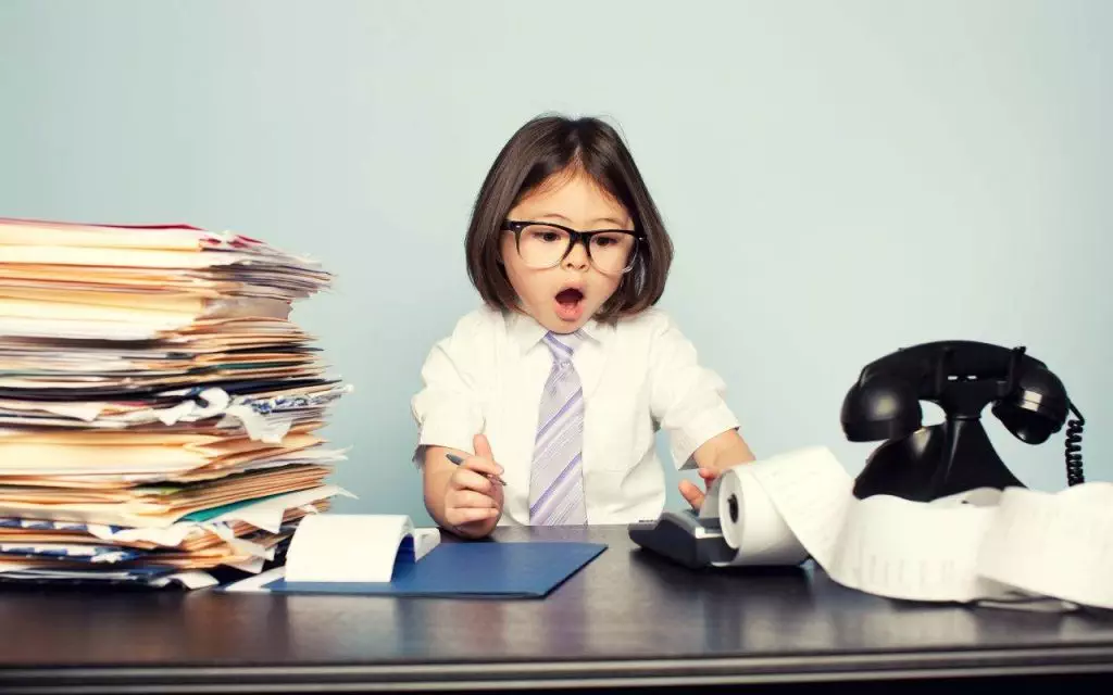 A little girl is sitting at a desk with a pile of papers, struggling to manage the accounting tasks for small businesses - top accounting pain points facing small businesses - not being prepared for unexpected costs.